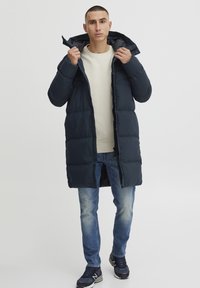 Mørk marineblå puffjakke med hette, med glidelås foran, store puffer og en jevnt tekstur. Båret over en lys genser og bukser.