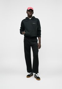 Joven con sudadera negra y pantalones negros, zapatillas en blanco y negro, y gorra de béisbol roja, de pie con una mano en el bolsillo.