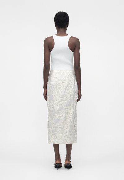 Γυναίκα που στέκεται με την πλάτη προς την κάμερα, φορώντας λευκό αμάνικο τοπ, μακριά κρεμ φούστα με σχέδια και μαύρα πέδιλα με τακούνι σε λευκό φόντο.