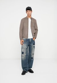 Chaqueta de ante marrón claro con cuello alto, combinada con jeans azules desgastados que tienen parches. El modelo lleva zapatos negros y una gorra.