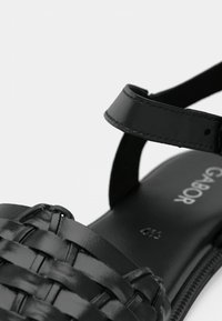 Μαύρο σανδάλι με πλεκτό επάνω μέρος από δέρμα και λουρί με λείο φινίρισμα. Η πάτος είναι μαύρη με διακριτικό λογότυπο.