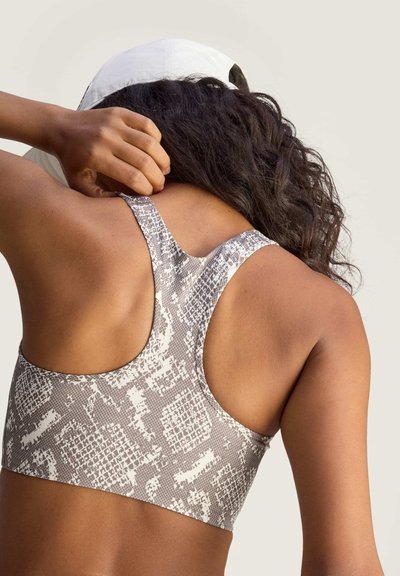 Vrouw met donker krullend haar, draagster van een grijs-witte sportbeha met racerback en patroon, die een witte pet afstelt tegen een beige achtergrond.