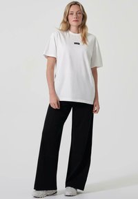 Bílé oversized tričko s krátkým rukávem a černou logovou nášivkou, spárované s černými širokými kalhotami a šedými teniskami. Jednoduchý a moderní design.