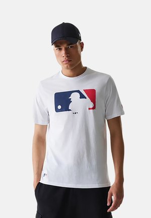 Junger Mann mit schwarzer Kappe, weißem T-Shirt mit blauem und rotem Baseball-Logo und schwarzer Shorts, steht mit den Händen in den Taschen.