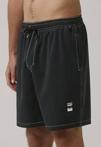 Pantaloni scurți de baie negri cu talie elastică și șnur, având o cusătură albă, buzunare laterale și un patch cu logo pe margine.