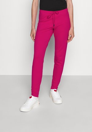 Personne portant un pantalon slim rose vif avec un cordon à la taille et des baskets blanches avec des détails en forme de cœur rouge.