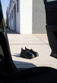 Par de zapatos Mary Jane negros de plataforma colocados en una acera iluminada por el sol, vistos desde el interior de una puerta de coche abierta en una calle de la ciudad.