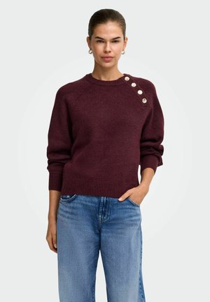 Jeune femme portant un pull bordeaux avec quatre boutons dorés sur l'épaule et un jean bleu, debout avec une main dans la poche.
