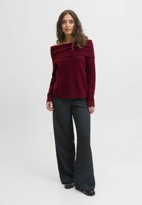 Vīnsarkanā džempera bez pleciem ar teksturētu ribveida apkakli, kombinēts ar vaļīgām melnām mirdzošām biksēm ar augstu jostasvietu un spīdīgiem apaviem.