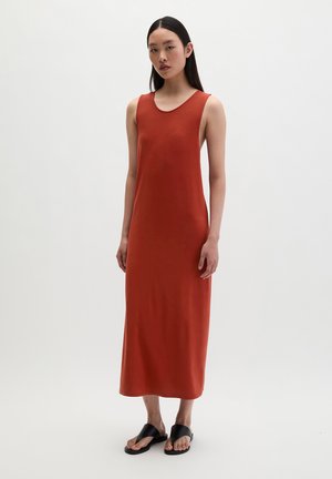 Frau mit langen dunklen Haaren, die ein ärmelloses rostfarbenes Midikleid und schwarze Sandalen trägt, vor einem schlichten hellen Hintergrund stehend.