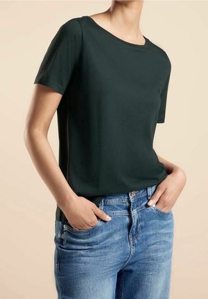 Personne portant un t-shirt noir uni à manches courtes et un jean en denim bleu, les mains partiellement dans les poches, sur fond beige.