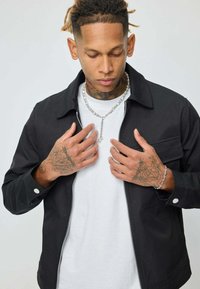 Jeune homme avec des tatouages sur les mains et le cou, portant une veste zippée noire, une chemise blanche, un collier en argent et un bracelet, qui ajuste sa veste.