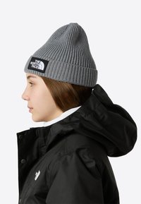 Γκρι πλεκτός σκούφος με ριμπ ύφανση και λογότυπο, φορεμένος με μαύρο μπουφάν με κουκούλα και λευκή εσωτερική στρώση, εμφανής από το πλάι.