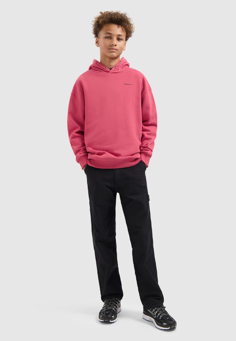 Tiener met handen in de zakken, gekleed in een roze hoodie, zwarte broek en zwarte sneakers, staand tegen een effen lichte achtergrond.