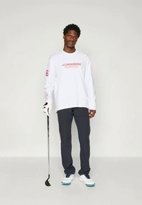 Langermet hvit skjorte med rosa og oransje tekst, sammen med mørke bukser. Modellen holder en golfklubbe, iført hvite og blå joggesko.