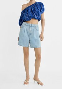 Top azul sin mangas con un patrón abstracto blanco; combinado con pantalones cortos de mezclilla azul claros; modelo lleva sandalias de tacón blancas.