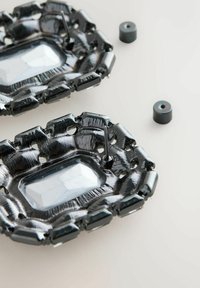 Schwarze rechteckige Ohrringe mit facettierten klaren Steinen in der Mitte und zwei schwarzen runden Rückseiten auf einer weißen Oberfläche.