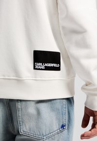 Felpa bianca con polsini a coste, dotata di una toppa rettangolare nera con la scritta "KARL LAGERFELD JEANS" in testo bianco. Visibili jeans azzurri chiari.