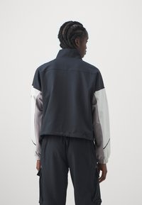 Sportjacke mit hohem Kragen; marineblauer Körper, weiße und graue Ärmel. Verfügt über elastische Bündchen und ein verkürztes Design. Glatte Textur.