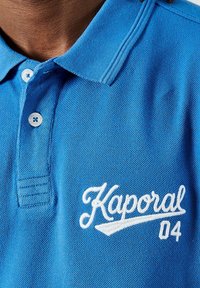 Blauwe polo gemaakt van textielstof, met een klassieke kraag en twee knopen. Witte geborduurde "Kaporal 04" logo aan de voorkant.