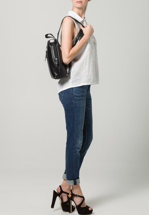 Schwarzer Leder-Rucksack, der über der Schulter getragen wird, mit verstellbaren Trägern und einem eleganten Design, kombiniert mit einem weißen Oberteil und blauen Jeans.