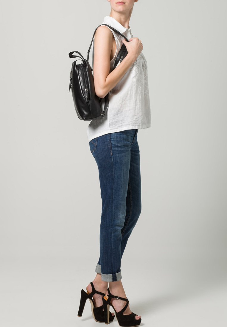 Un sac à dos en cuir noir porté sur l'épaule, avec des bretelles ajustables et un design élégant, associé à un haut blanc et un jean bleu.