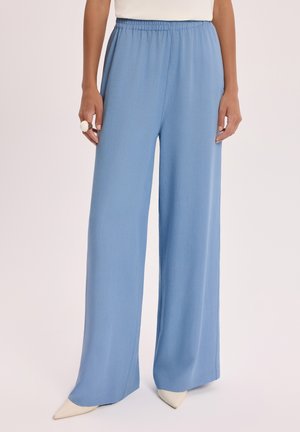 Kobieta w jasnoniebieskich spodniach o szerokich nogawkach z elastycznym pasem i białych, spiczastych butach, stojąca na tle jednolitego tła.