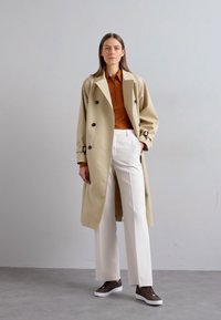 Béžový trenčkot s veľkými čiernymi gombíkmi, cez hnedú košeľu a biele široké nohavice. Hnedé tenisky dopĺňajú outfit.