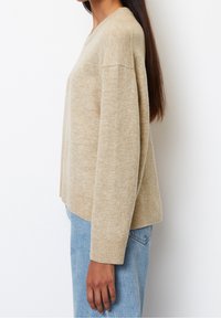 Pull beige en maille à coupe décontractée, avec poignets côtelés et col rond. Porté avec un jean taille haute bleu clair, vue de côté.