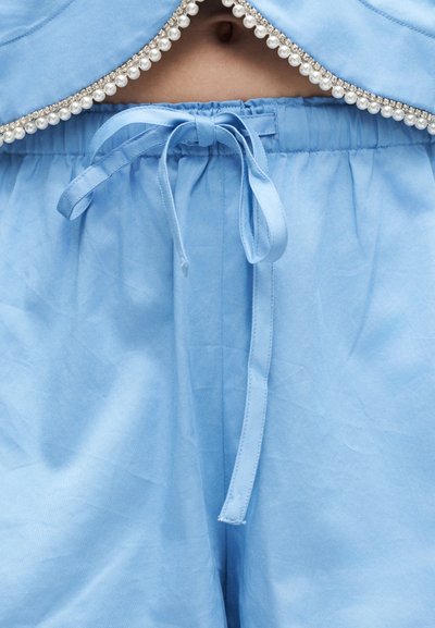 Pantalon en tissu bleu clair avec une ceinture à cordon noué, et un haut assorti bordé de perles et de strass.