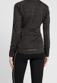 Mörkgrå atletisk långärmad topp med en strukturerad tyg, utrustad med en ficka med dragkedja på baksidan och reflekterande detaljer, matchad med svarta tights.