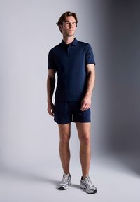 Tamsiai mėlynas trumpomis rankovėmis polo marškinėliai siauru siluetu, derinami su atitinkančiais šortais. Modelis dėvi šviesiai pilkus sportinius batelius. Sklandus tekstilės paviršius.