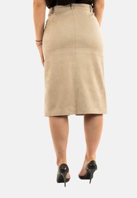 Femme portant une jupe beige jusqu'aux genoux, un haut noir et des chaussures à talons noirs, se tenant avec les mains détendues le long du corps.