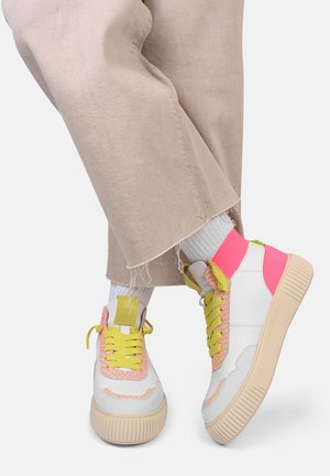 Sneakers bianchi con accenti neon rosa e giallo, tessuto testurizzato, suola prominente e lacci a fantasia; abbinati a pantaloni beige a gamba larga.