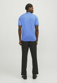 Ljusblå kortärmad polo med en jämn textur, i kombination med svarta raka byxor och svarta skor, sedd bakifrån.