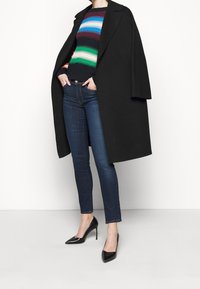 Abrigo negro, suéter de punto a rayas en azul, verde y rojo, jeans skinny azul oscuro y tacones negros de punta. Texturas suaves y líneas limpias.