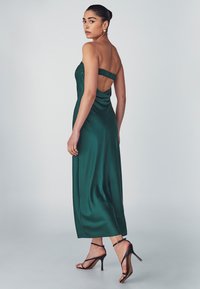 Abito in satin verde smeraldo con design senza spalline, arricciatura sul retro e silhouette fluida; abbinato a sandali neri con tacco alto e cinturini.