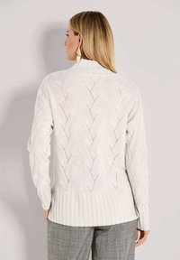 Frau mit glattem, blonden Haar trägt einen cremefarbenen Pullover aus Strick mit strukturiertem Zickzackmuster und grauen karierten Hosen, die von der Kamera wegblickt.