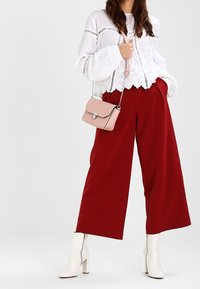 Bílá krajková halenka s vroubkovaným lemem, kombinovaná s červenými kalhotami s širokými nohavicemi a bílými kotníkovými botami. Součástí je malá růžová kabelka přes rameno.