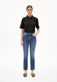 Zwarte polo met korte mouwen en een ritsdetails, gecombineerd met lichtblauwe high-waisted rechte jeans. Zwarte instap schoenen maken de outfit compleet.