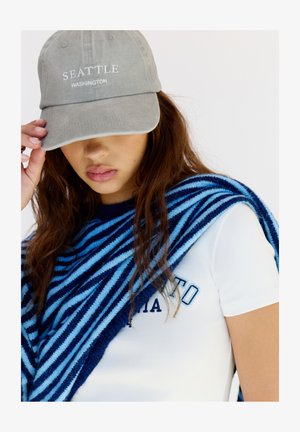 Cappello di cotone grigio con ricamo "SEATTLE WASHINGTON". Sciarpa in maglia a righe blu e navy drappeggiata su una t-shirt bianca con la scritta "CALIFORNIA".