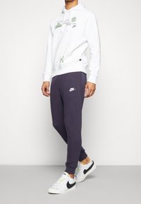 Vit hoodie med grön grafisk design, frontficka; ihop med mörklila sweatpants med en vit Nike-logotyp; vita och svarta sneakers.