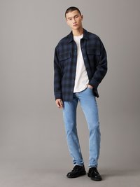 Camisa de cuadros en azul marino y negro sobre una camiseta blanca, combinada con vaqueros de mezclilla azul claro y zapatos negros. Textura suave y corte holgado.