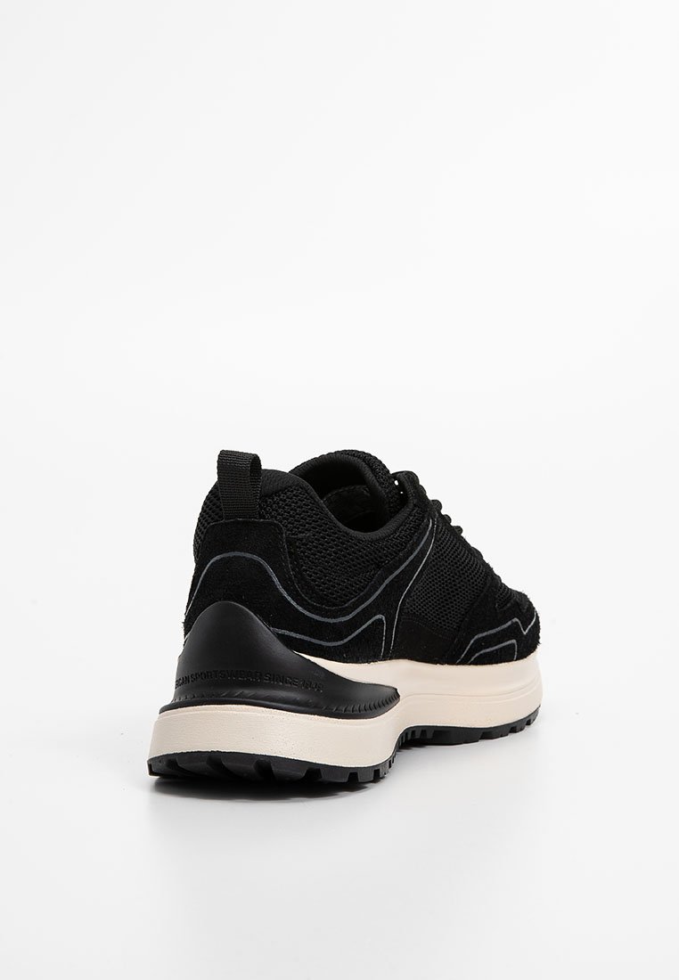 Baskets noires en maille et daim avec une épaisse semelle blanche, un capuchon en caoutchouc au talon et une languette à l'arrière, vues de l'angle arrière sur fond blanc.