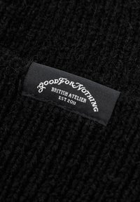 Tessuto a maglia nero con una superficie strutturata. Un'etichetta cucita presenta un testo bianco che recita "Good For Nothing British Atelier Est 2011."