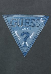 Parche de logo de denim en forma triangular, presenta una tela azul con texto bordado sobre un fondo oscuro. Se aprecian acentos desgastados.