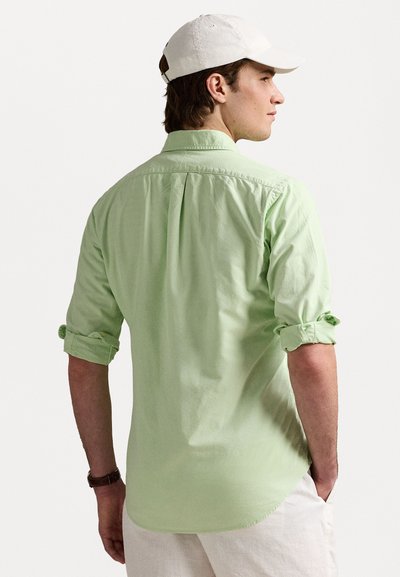 Camisa de botón de color verde claro hecha de algodón, con mangas enrolladas, un pliegue en la espalda y un dobladillo curvado, combinada con pantalones de color claro.