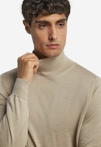 Beżowy sweter z golfem wykonany z miękkiego, drobno splotowego materiału. Dopasowany krój z prążkowanymi mankietami i gładką teksturą. Model trzyma kołnierz.