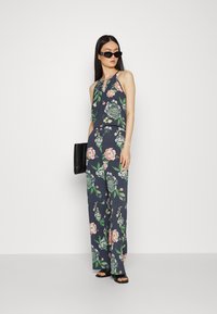 Macacão floral em tecido azul marinho, com decote halter, pernas largas e padrões distintos de flores verdes e rosas. Acessorizado com óculos de sol pretos.