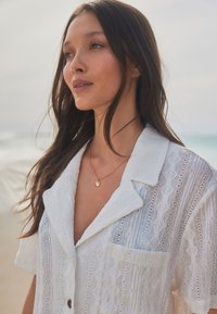Chemise blanche texturée à manches courtes avec un motif en losange et une poche, associée à un délicat collier en or, sur un fond de plage.